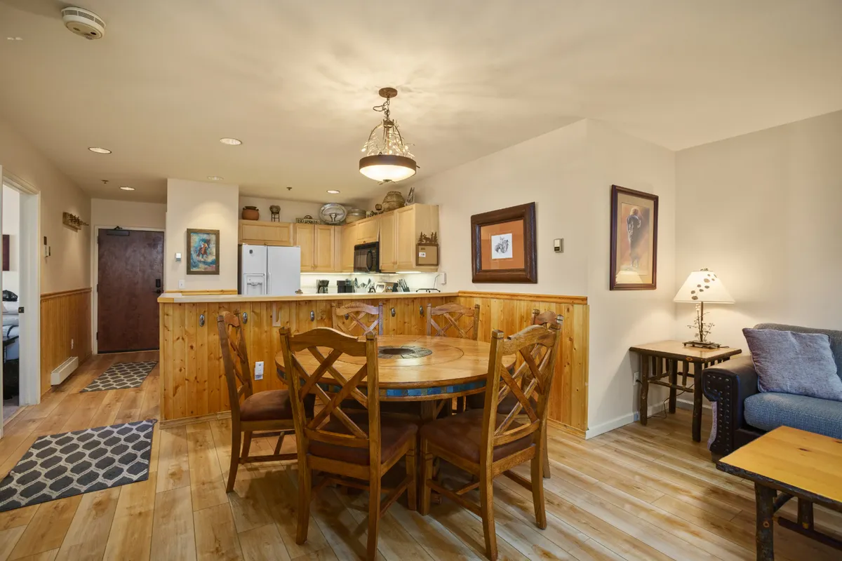 A wide interior shot showing an open-concept space with a round dining table set for a meal, comfortable sofas arranged around a living area, and a glimpse of the kitchen in the background. Natural light pours in, enhancing the inviting atmosphere, while the room is set up for both family meals and social gatherings.