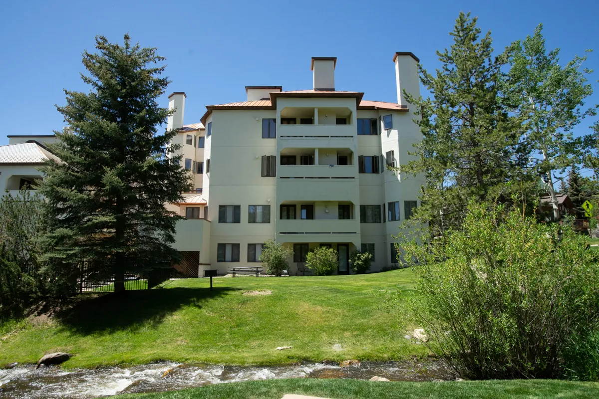 This view highlights the building’s facade, a lush green lawn, towering trees, and a flowing stream in the foreground. The scene evokes tranquility and the close connection to nature, while also showing the proximity of the condo to outdoor village features.
