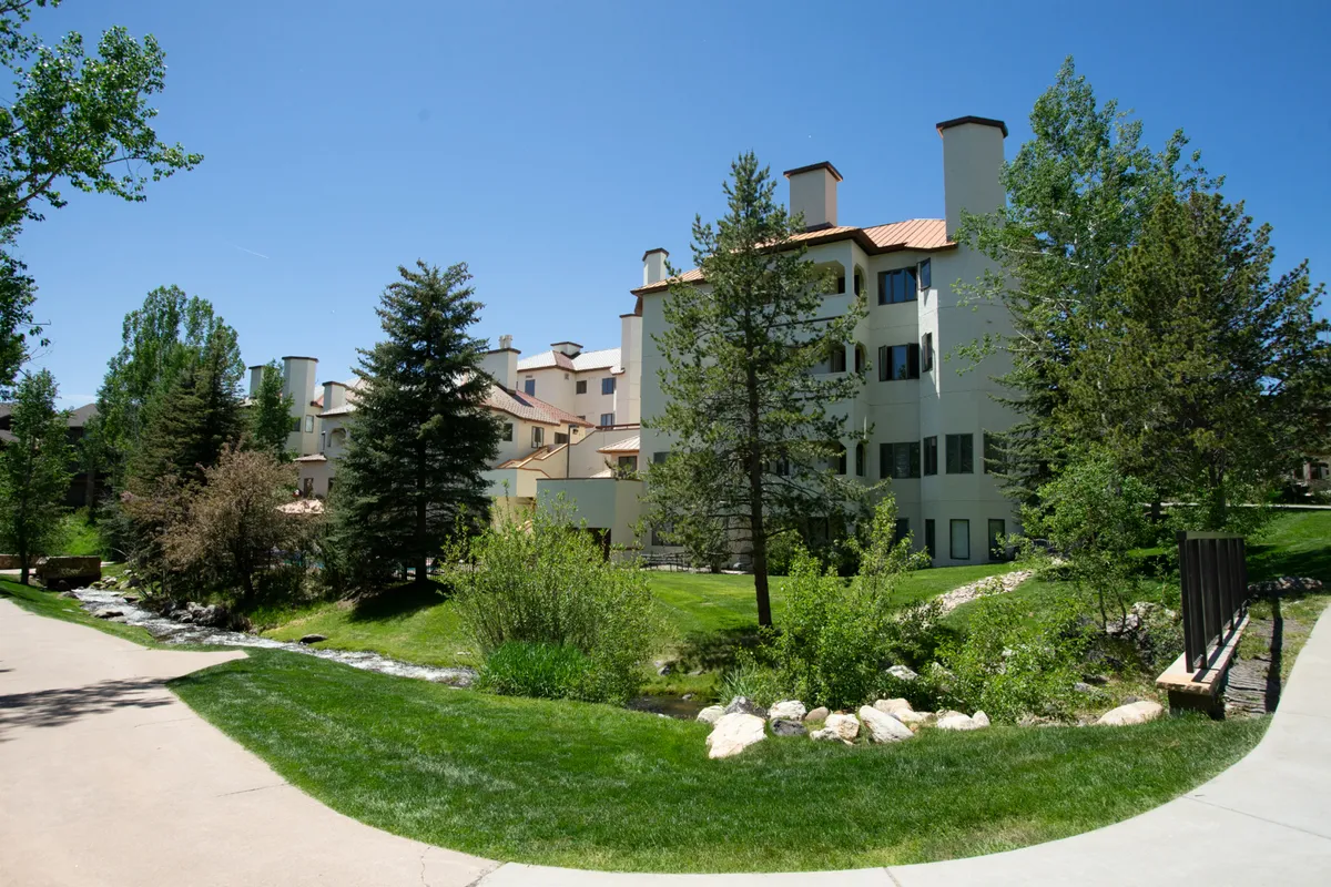 A peaceful scene showing a building facade bordered by lush greenery and a picturesque flowing stream, with a winding pathway inviting exploration. The landscape feels both lively and tranquil, suggesting year-round enjoyment.