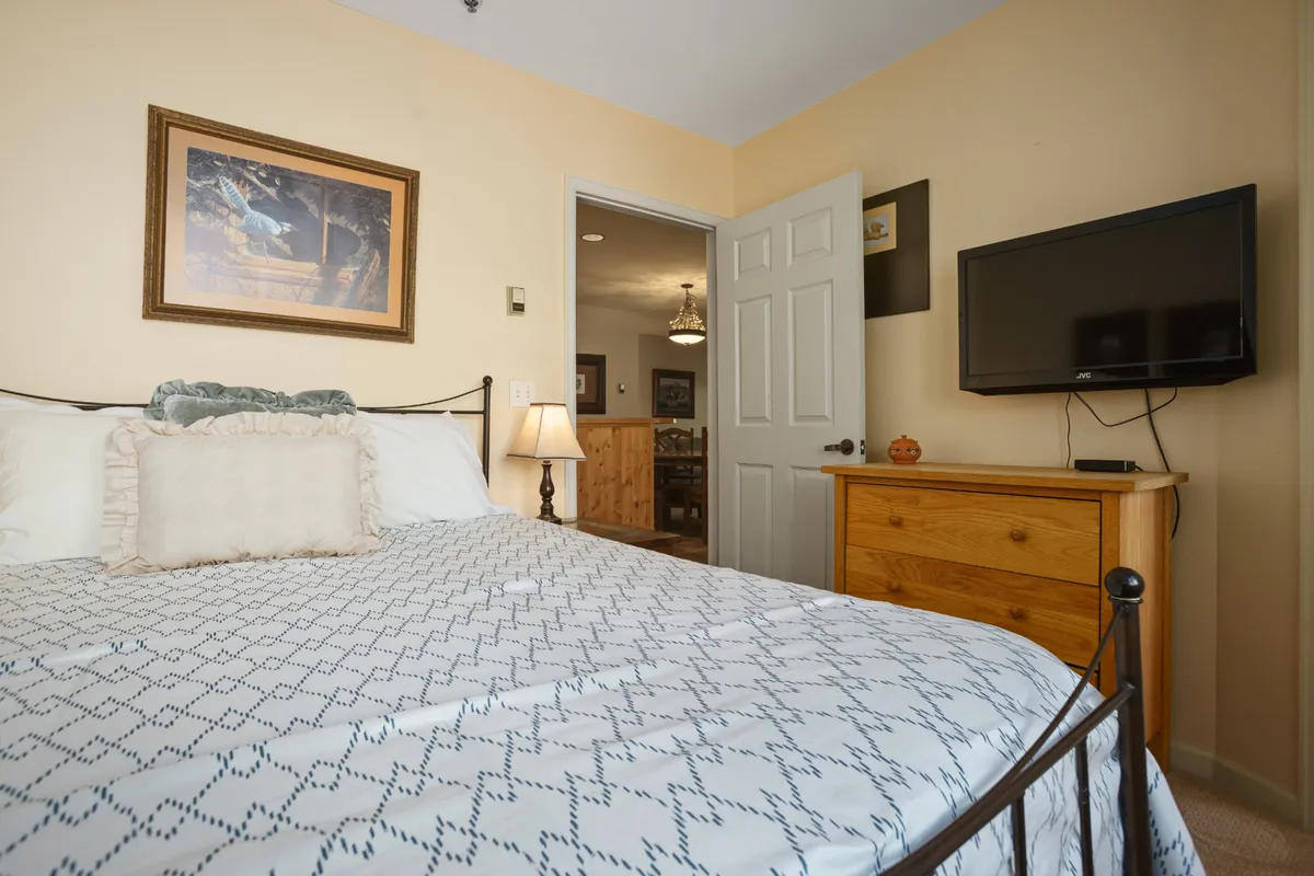 A welcoming bedroom featuring a queen-sized bed with cozy patterned bedding, a wooden dresser with a TV atop, and framed artwork on the wall. The space is uncluttered and homey, reflecting a relaxing environment for unwinding or movie nights in bed.