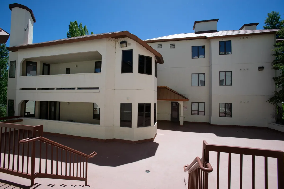 The architectural design of the building is showcased, with a staircase leading up to the welcoming entrance and lush greenery around. The area appears social and accessible, perfect for gatherings before heading out or returning from town activities.