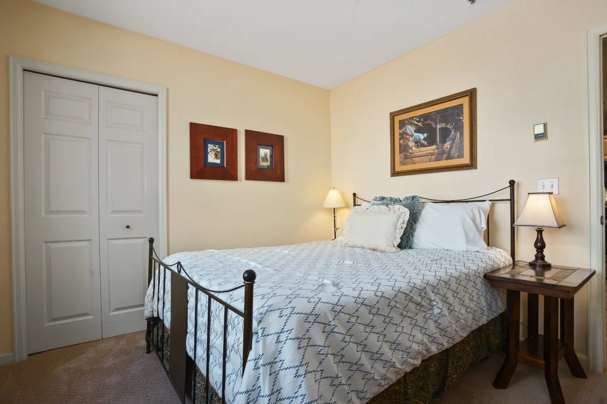 A master bedroom with a plush bed, neatly made with inviting linens, sits beside a nightstand and lamp. Wall art and gentle lighting contribute to a serene, restful ambiance, making the space feel calm and comfortable.