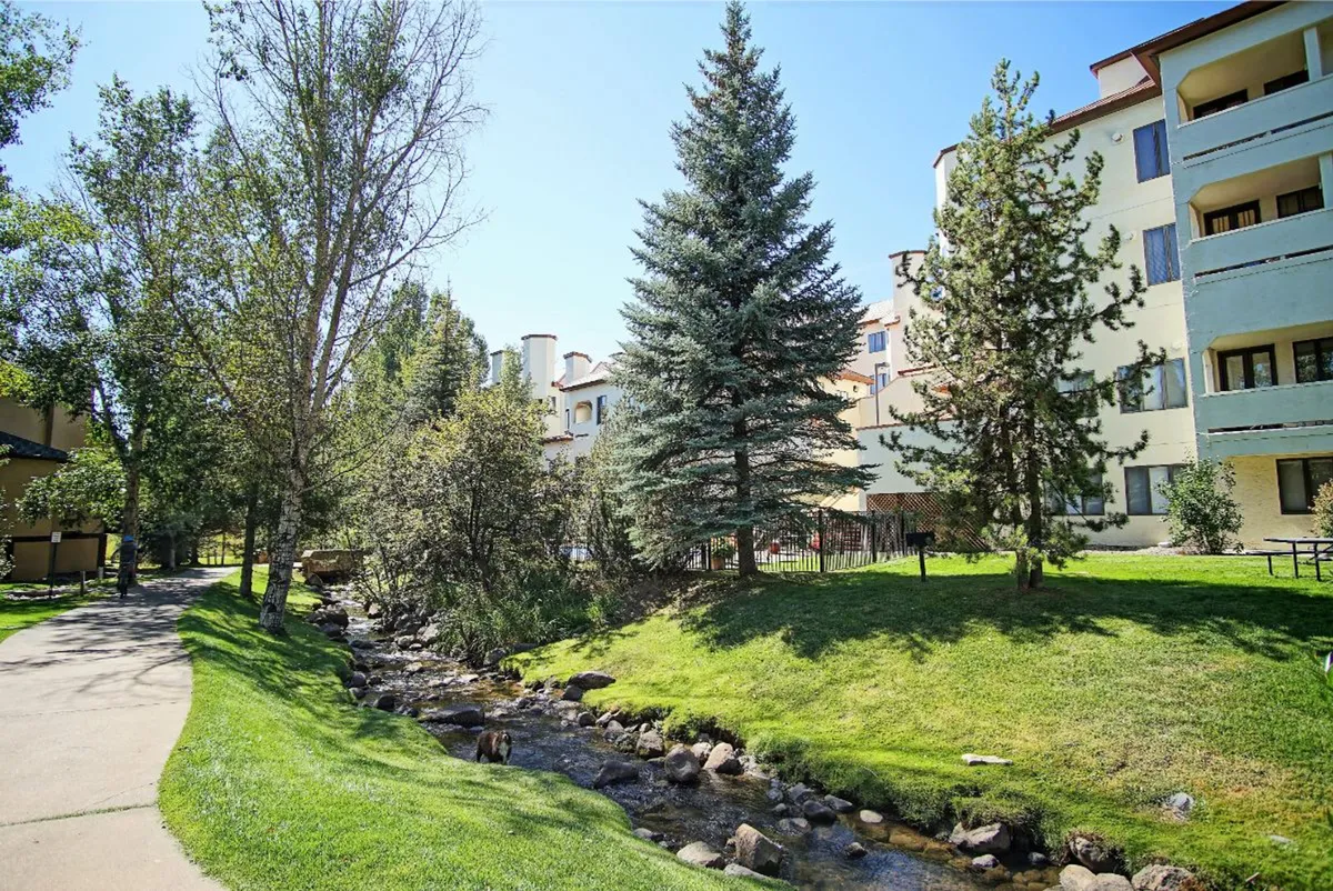An inviting creekside scene featuring a natural stream with lush trees lining the banks, a building in the background, and open grassy areas ideal for relaxing or strolling. The tranquil setting captures the peaceful, romantic vibe of the location and the proximity to nature's beauty.