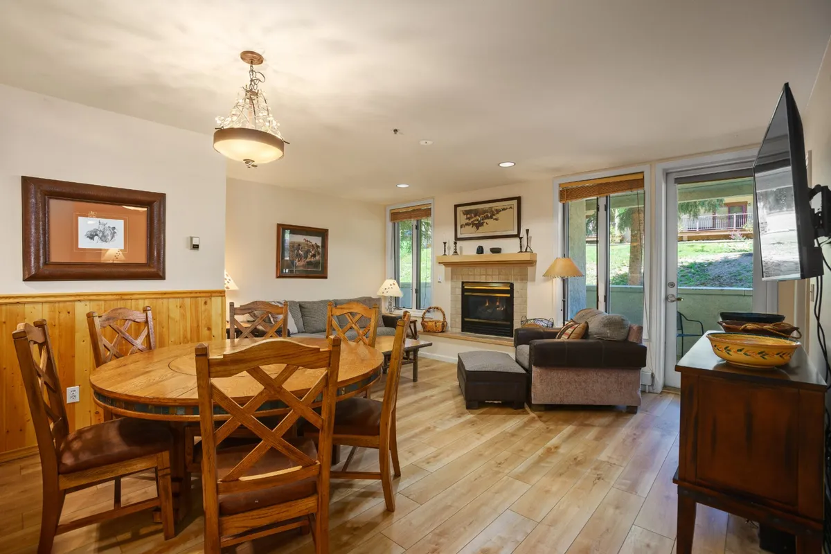 A cozy interior featuring a round dining table positioned near a warm fireplace, with large windows letting in sunlight. The space is clearly used for both intimate dinners and group game nights, with the fire providing a welcoming focal point.