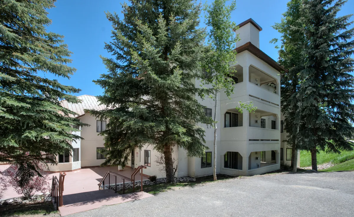 This image showcases the three-story building exterior surrounded by lush trees, with visible entrance stairs leading up to the main door. The balconies hint at the inviting living spaces inside, and the natural setting emphasizes the mountain village atmosphere.