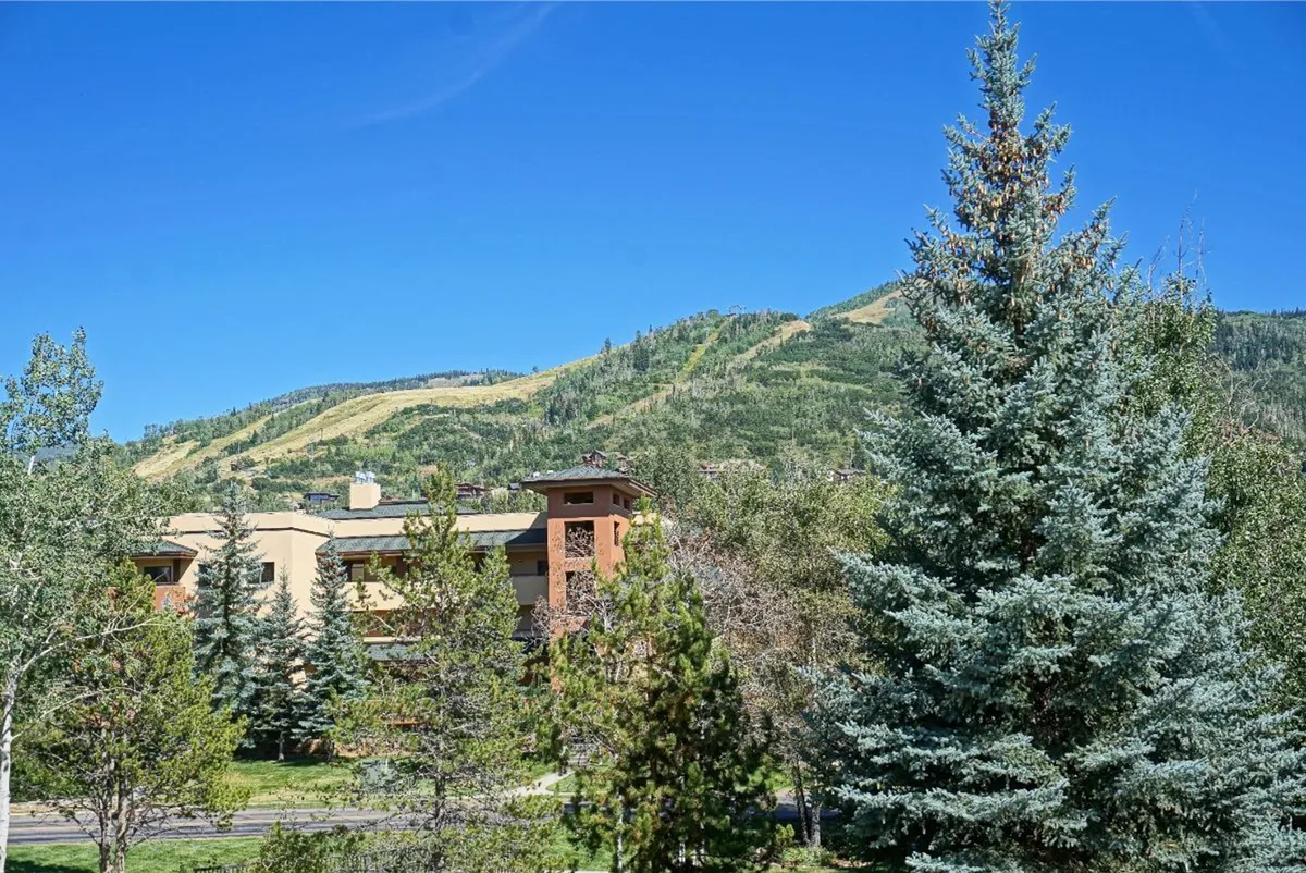 The image captures a scenic mountain backdrop beyond the condo building, surrounded by green trees. The perspective emphasizes the property’s mountain village setting and the proximity to hiking and nature trails.